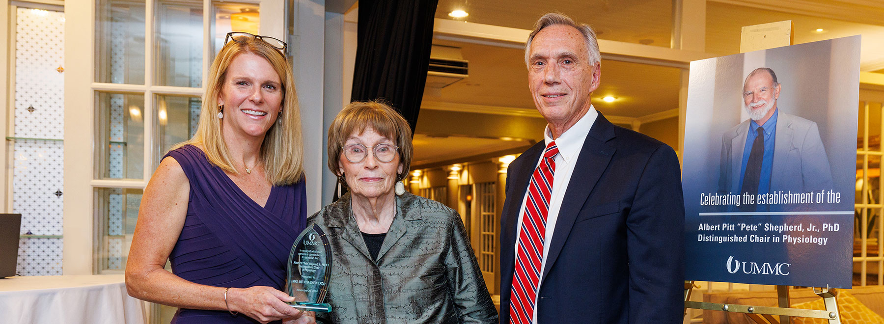 From left, Dr. Genevieve Shepherd Ali, Melissa Shepherd and Dr. John Hall present an award to Mrs. Shepherd recognizing her contribution to the Albert Pitt "Pete" Shepherd Jr., PhD Distinguished Chair in Physiology. To the right is a large display board featuring a photograph of Pete Shepherd and text that reads: “Celebrating the establishment of the Albert Pitt ‘Pete’ Shepherd, Jr., PhD Distinguished Chair in Physiology” along with the UMMC logo.